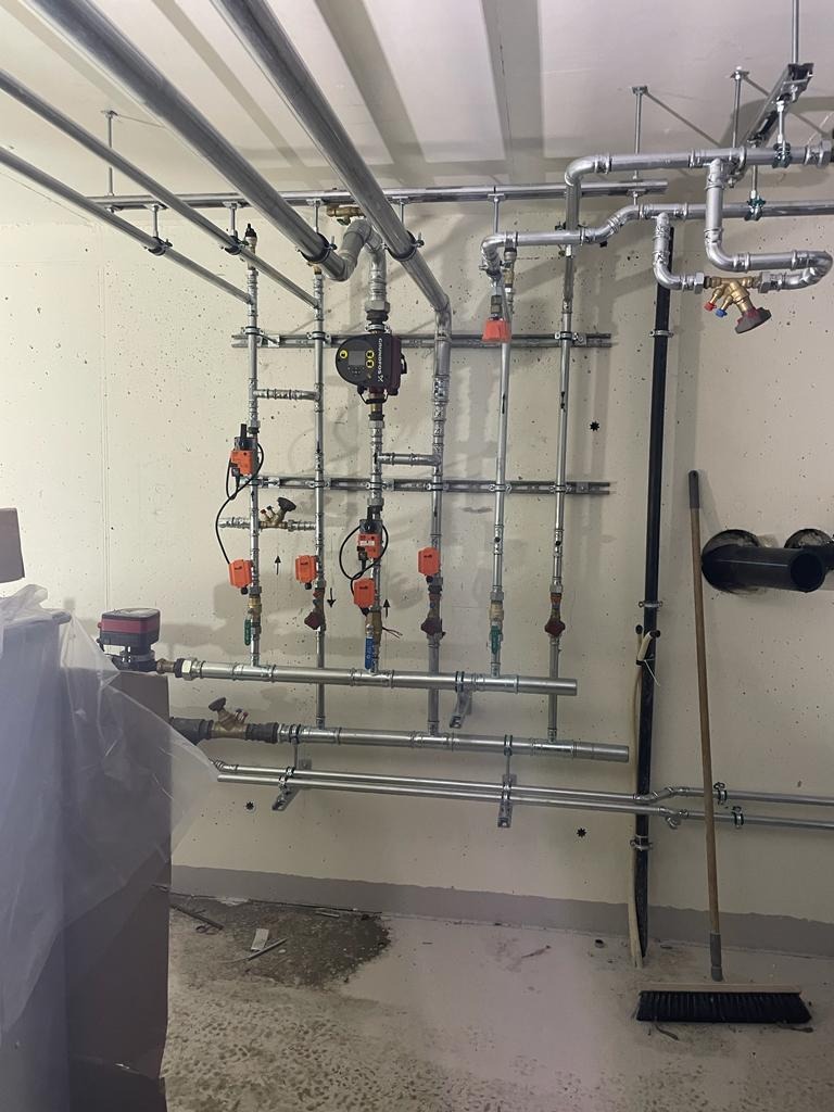 Złożona instalacja grzewcza z rurami, zaworami i pompą obiegową. Widoczne elementy mocujące do ściany i sufitu. Ujęcie z perspektywy podłogi, widoczny fragment kotła.