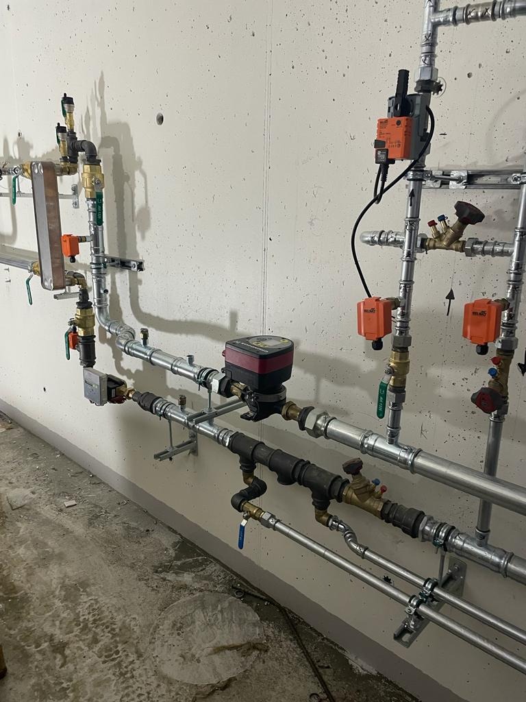 Złożona instalacja hydrauliczna z rurami, zaworami i czujnikami na szarej ścianie. Widoczne elementy grzewcze i wodne, całość tworzy skomplikowany system.