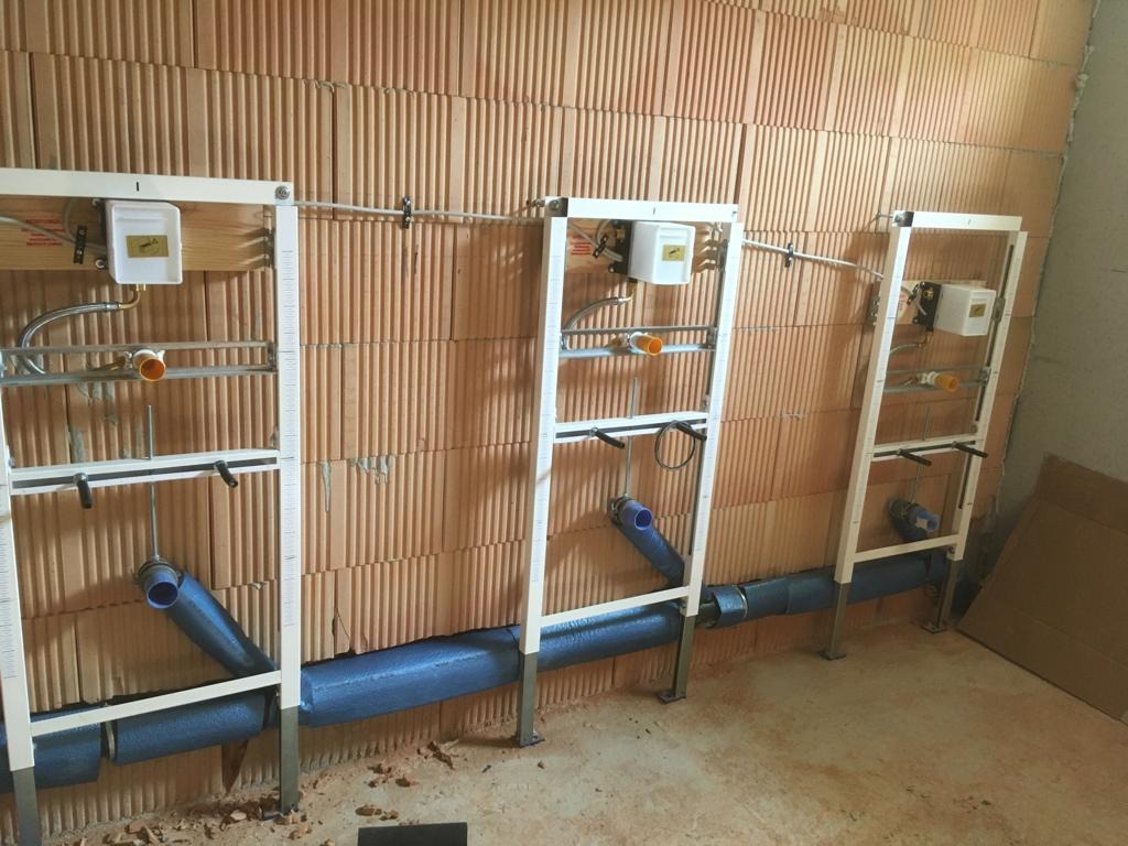 Instalacja wodno-kanalizacyjna w trakcie montażu: rury w izolacji, białe stelaże podtynkowe, ceglana ściana. Widoczne elementy przyłączeniowe i skrzynki.