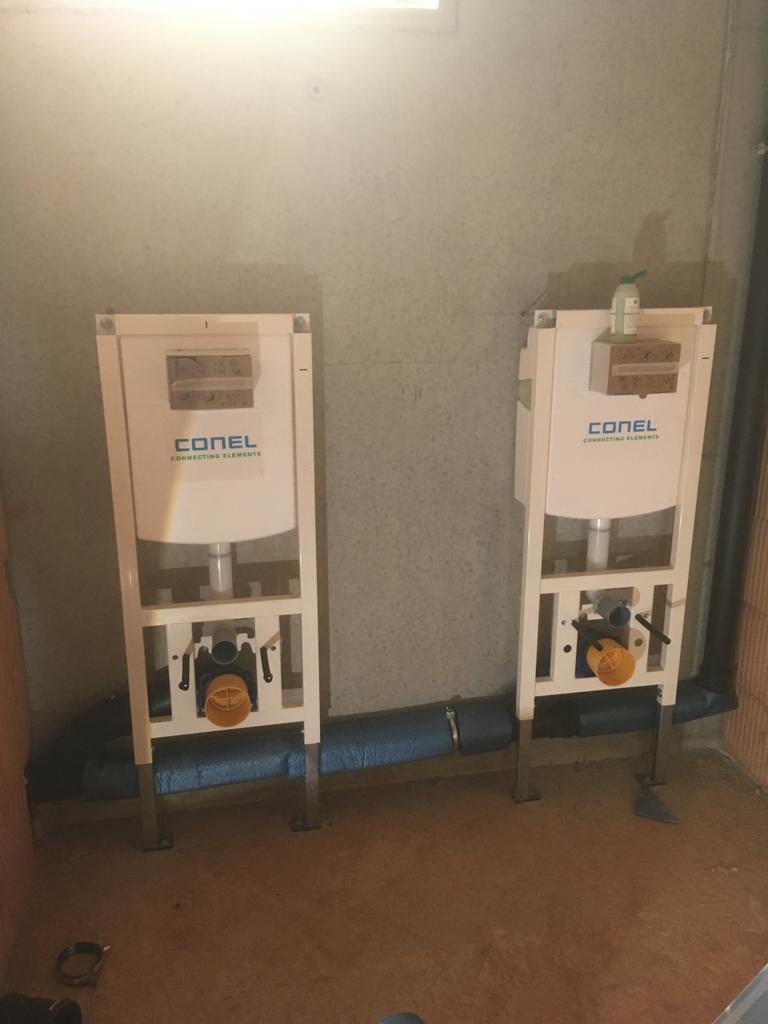 Dwa moduły WC podtynkowego marki Conel, połączone niebieską rurą kanalizacyjną, zamontowane przy szarej ścianie. Widoczne elementy instalacji i mocowania.