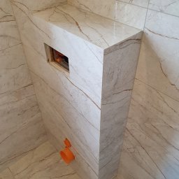 Perfect Design Grzegorz Siudak - Obudowa stelaża WC z płytek imitujących marmur, z widocznymi pomarańczowymi elementami instalacji. Ułożone płytki podłogowe w tym samym stylu.