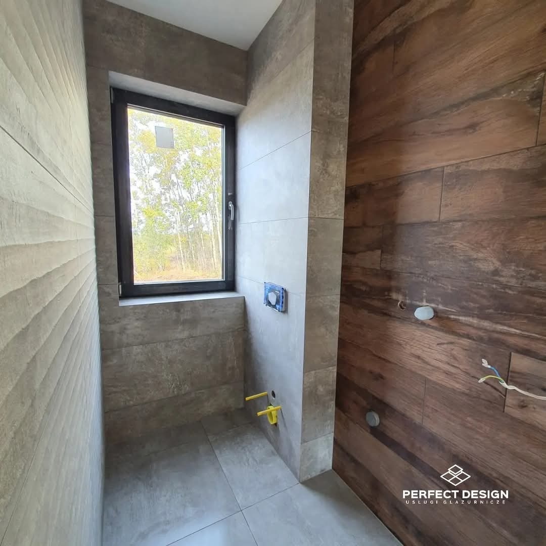 Nowoczesna łazienka z oknem, wykończona płytkami imitującymi drewno i beton. Widoczne przyłącza wodne i częściowo ułożone płytki. Minimalistyczny design.