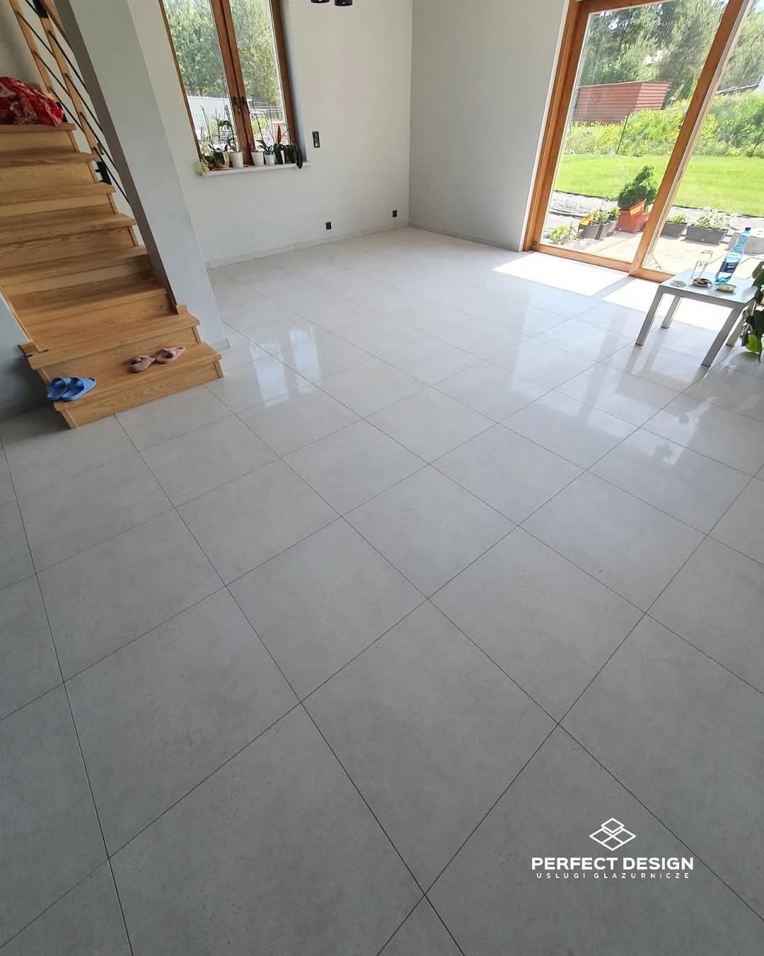 Jasne, błyszczące płytki podłogowe ułożone w salonie z drewnianymi schodami i dużym oknem w Siemonii. Widoczny fragment logo firmy glazurniczej.