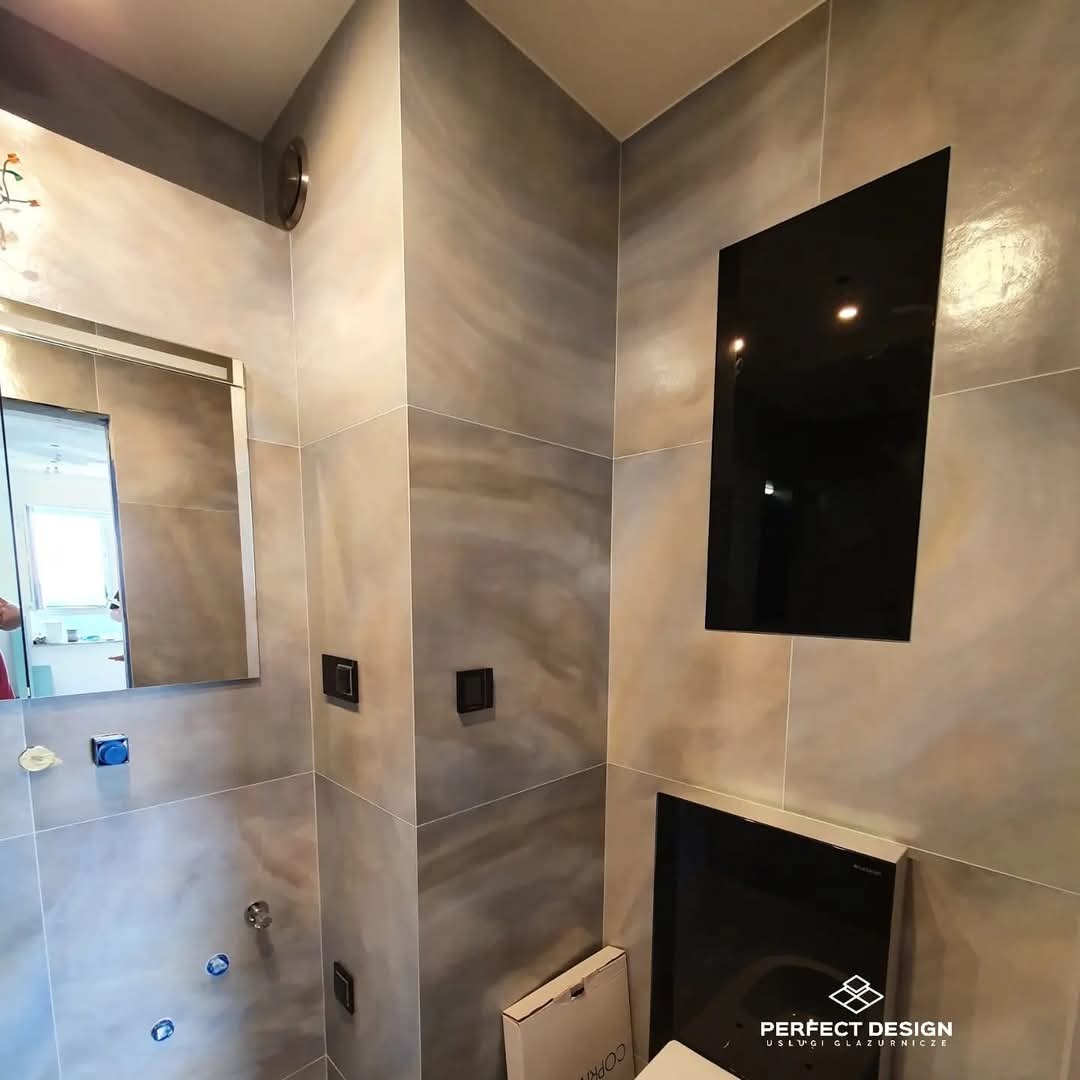Wykończona łazienka z szarymi płytkami imitującymi kamień, czarną zabudową i lustrem. Widoczne detale: bateria, włączniki i logo firmy glazurniczej.