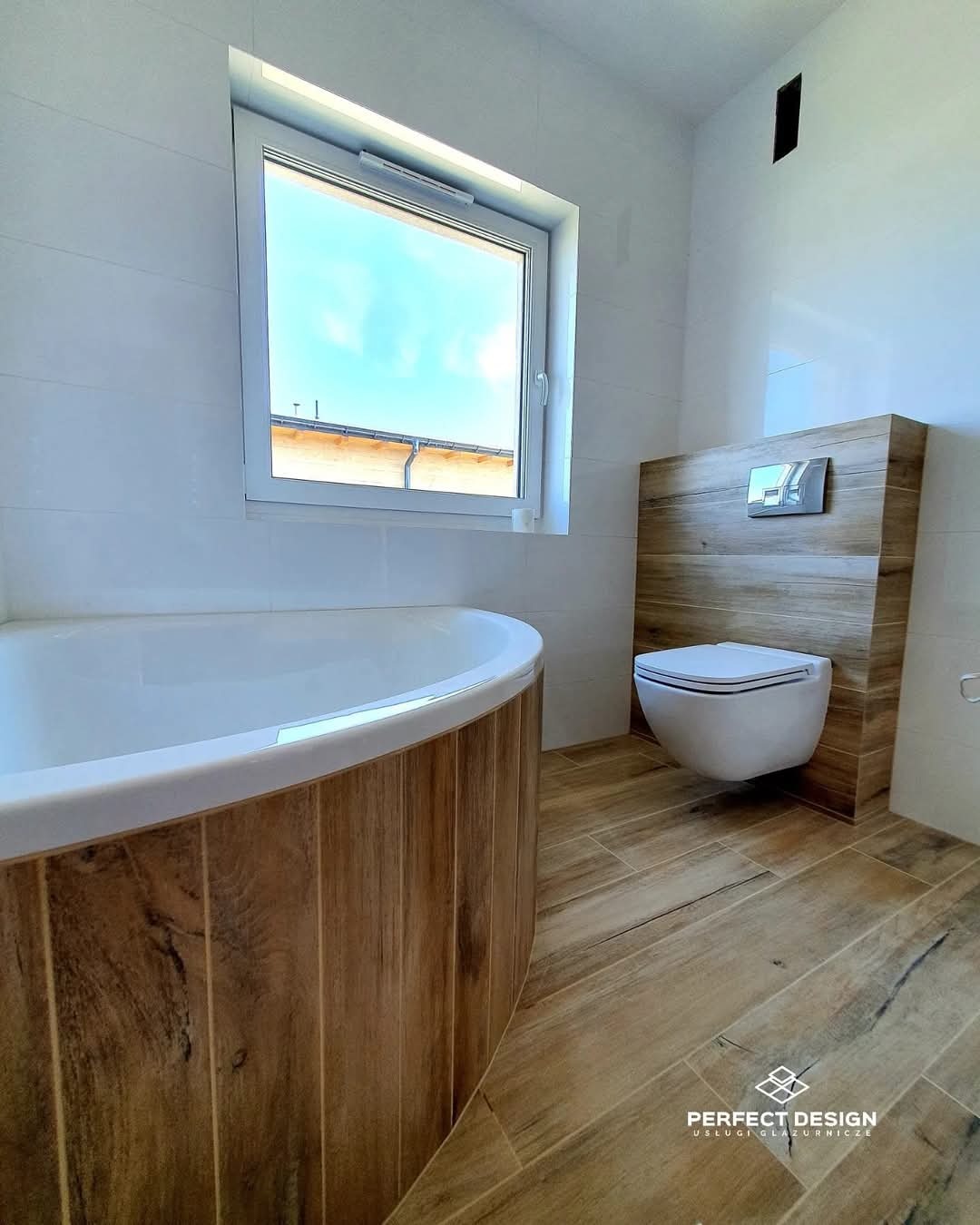 Nowoczesna łazienka z wanną obudowaną drewnem i podwieszaną toaletą. Płytki imitujące drewno na podłodze. Minimalistyczny styl, jasne kolory, okno z widokiem na niebo.