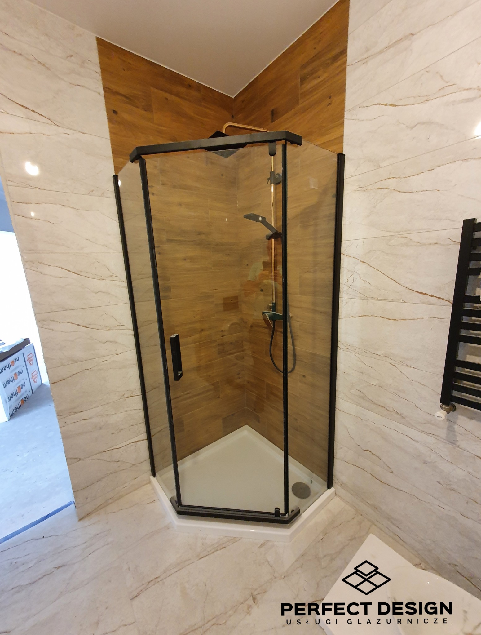 Kabina prysznicowa z czarnymi profilami w narożnej łazience, wykończonej płytkami imitującymi drewno i marmur. Widoczny grzejnik drabinkowy.