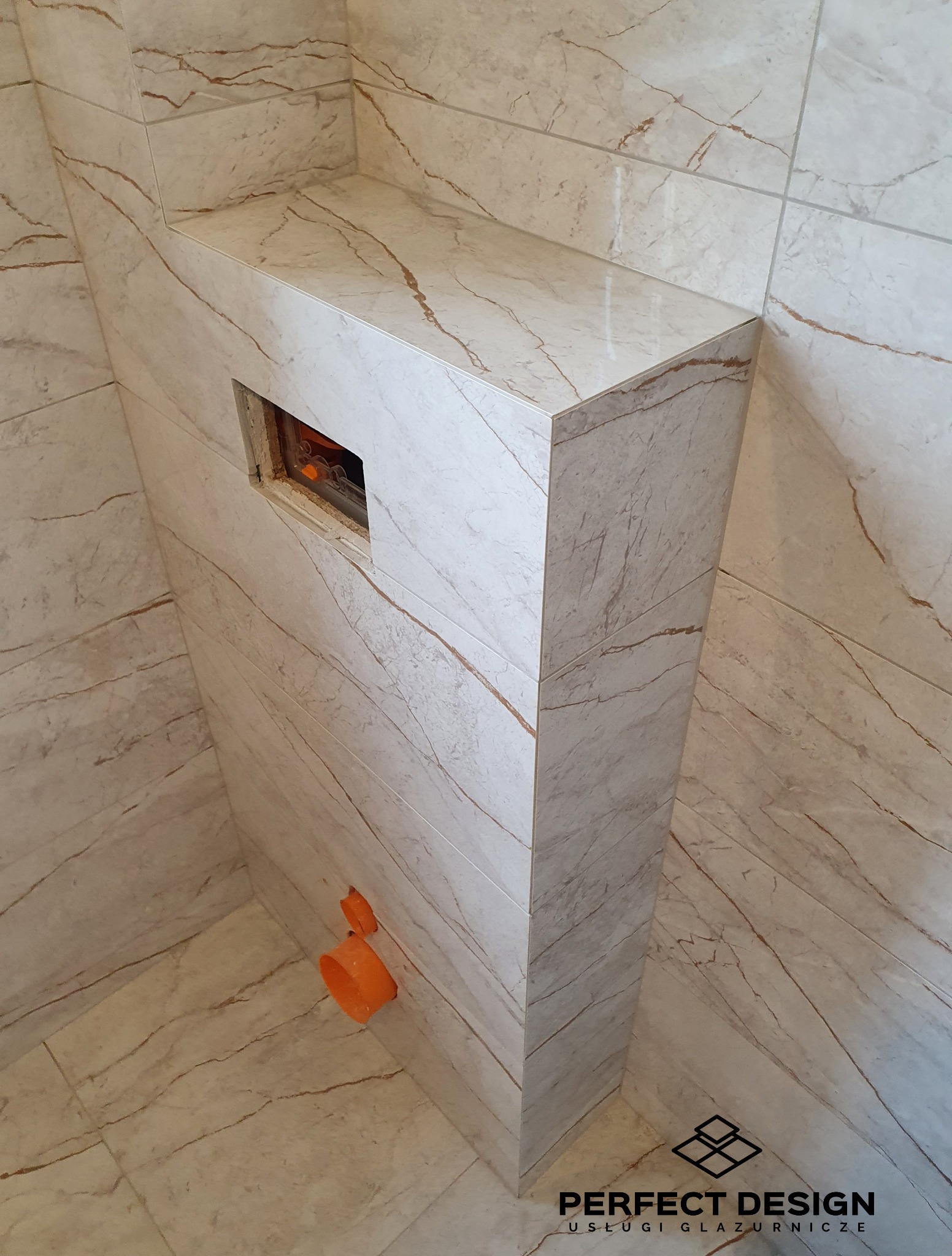 Obudowa stelaża WC z płytek imitujących marmur, z widocznymi pomarańczowymi elementami instalacji. Ułożone płytki podłogowe w tym samym stylu.