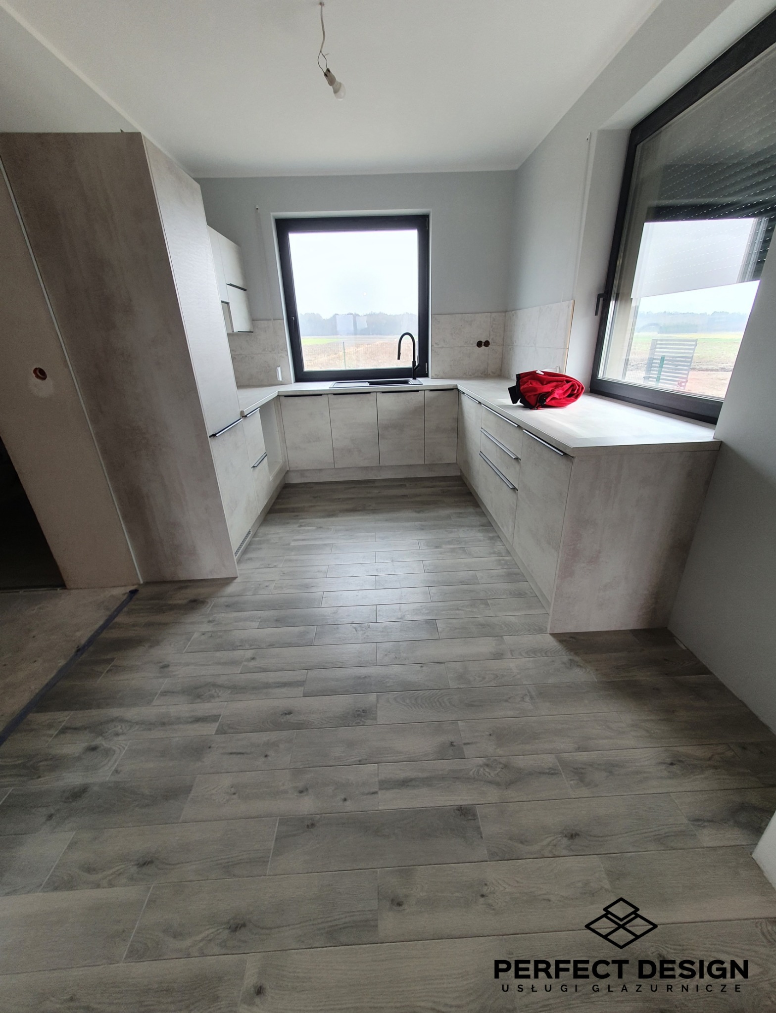 Jasna kuchnia w stylu nowoczesnym z drewnopodobną podłogą i czarną armaturą. Widoczny fragment niezabudowanej ściany.