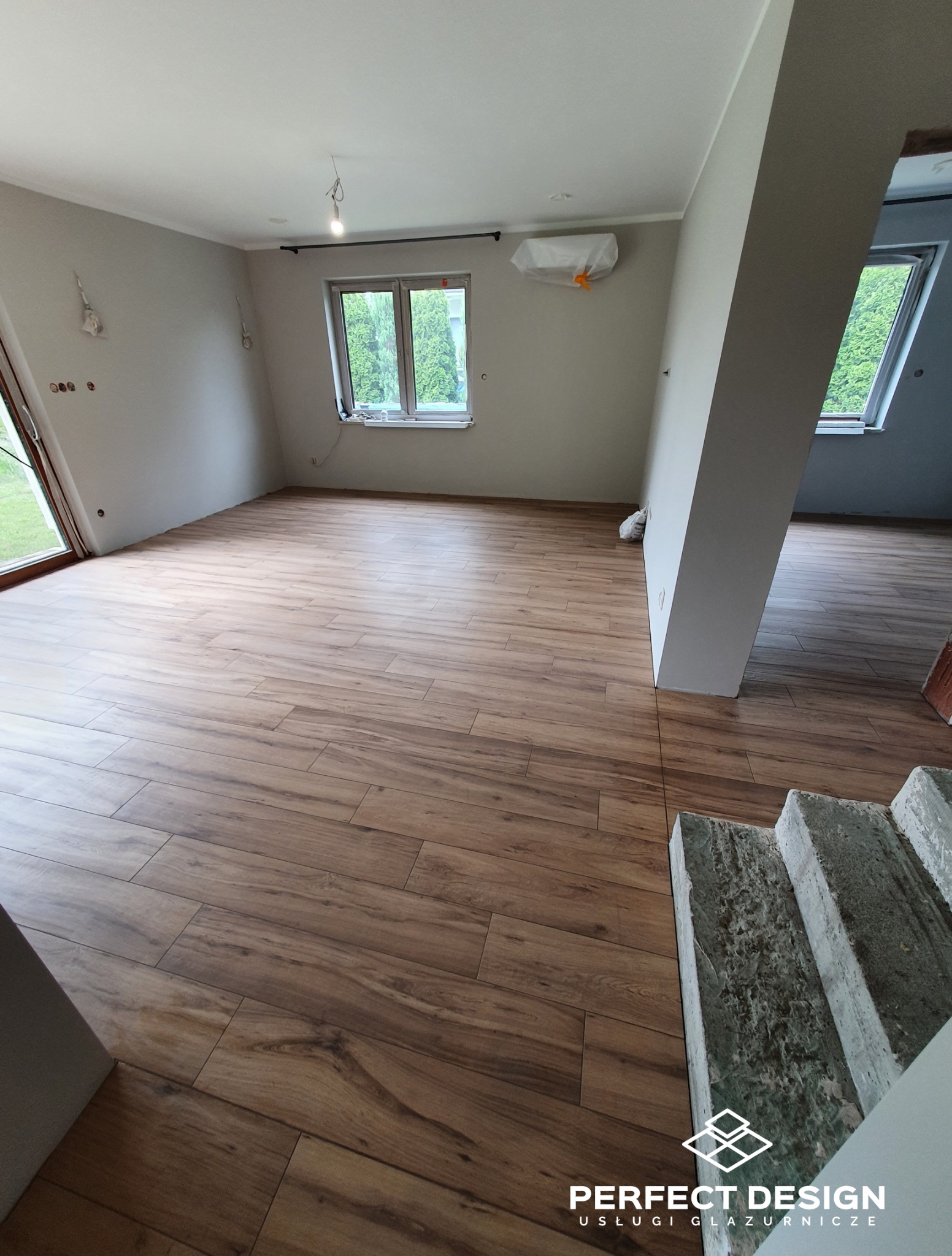 Wykończona podłoga z paneli imitujących drewno w jasnym wnętrzu. Widoczne okno, ściany przygotowane do malowania i surowe schody. Częściowo widoczny logo firmy.