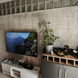 DECOROOM ARKADIUSZ RATAJCZAK - Ściana w salonie z efektem betonu architektonicznego, podkreślona dekoracyjnymi szklanymi pustakami. Telewizor i meble dopełniają nowoczesny wystrój.