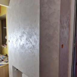 DECOROOM ARKADIUSZ RATAJCZAK - Ściana z dekoracyjną, srebrzystą gładzią wnęki, z widocznym kablem na suficie i wiadrem farby na podłodze. Wykończenie wnętrza z efektem połysku.