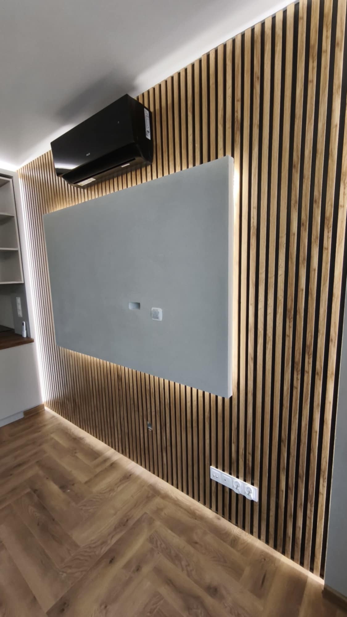 Ściana z drewnianych lameli z podświetleniem LED, szara płyta na TV, parkiet w jodełkę. Nowoczesny design wnętrza z klimatyzacją.