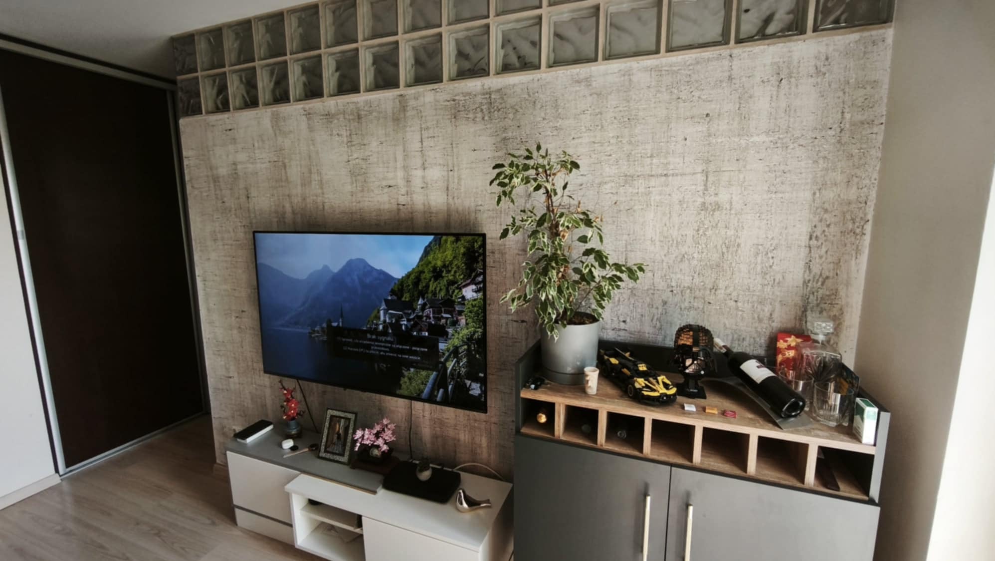 Ściana w salonie z efektem betonu architektonicznego, podkreślona dekoracyjnymi szklanymi pustakami. Telewizor i meble dopełniają nowoczesny wystrój.