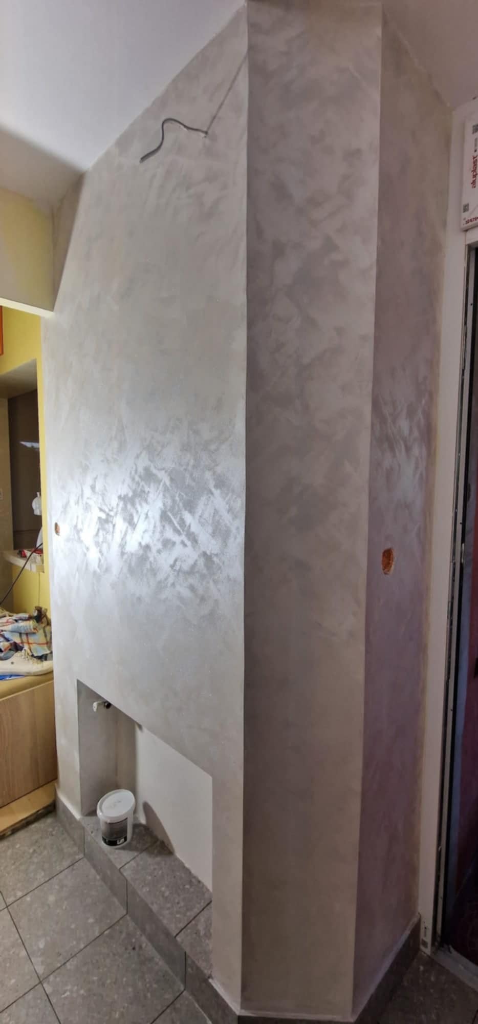 Ściana z dekoracyjną, srebrzystą gładzią wnęki, z widocznym kablem na suficie i wiadrem farby na podłodze. Wykończenie wnętrza z efektem połysku.