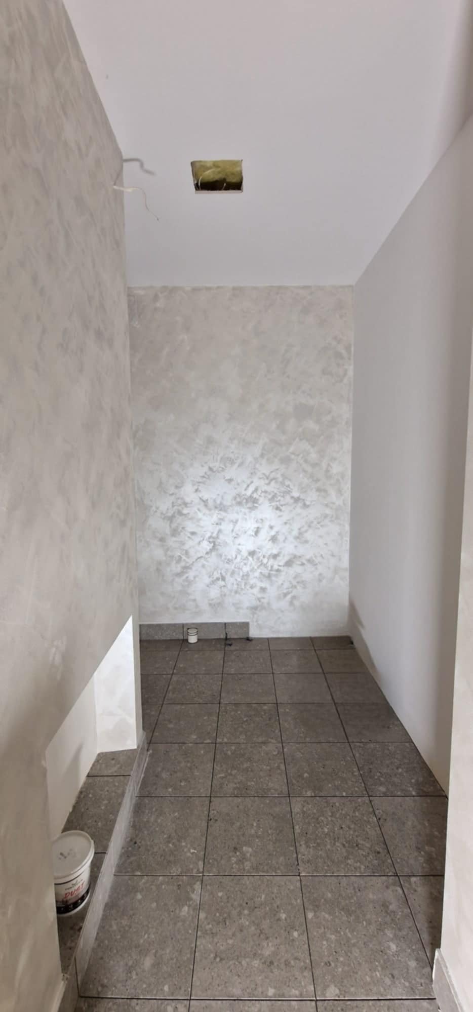 Wnętrze korytarza po gładzi dekoracyjnej w odcieniach szarości. Widoczna puszka elektryczna w suficie i wiadro farby przy ścianie. Podłoga wyłożona szarymi płytkami.