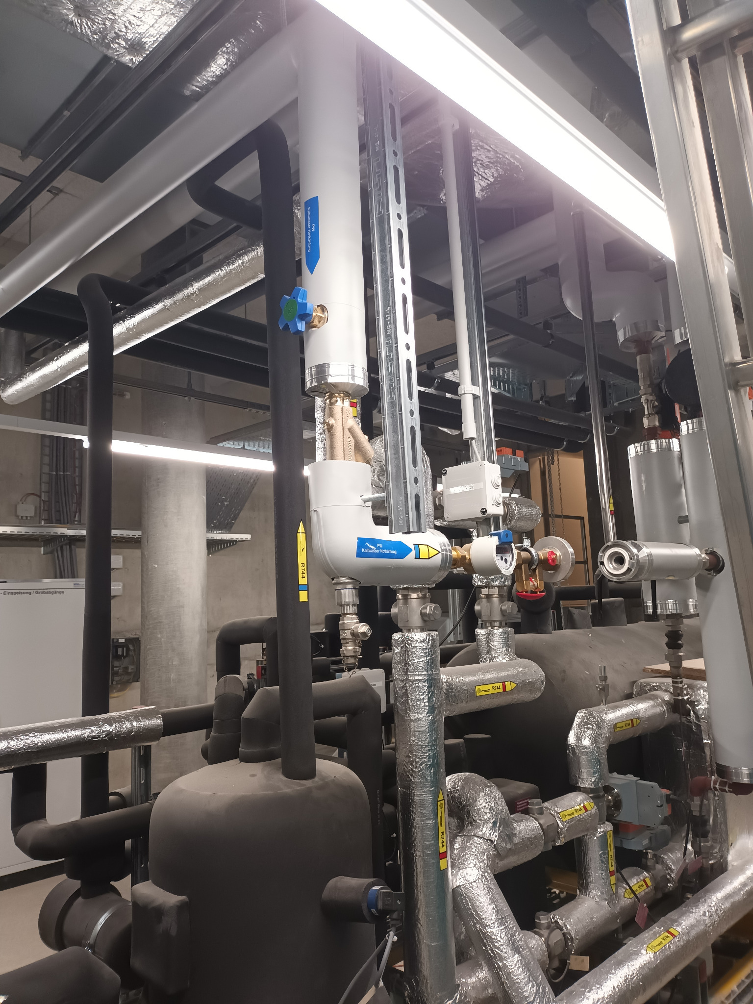 Złożona instalacja przemysłowa z rurami, zaworami i izolacją. Widoczne oznaczenia na rurach i elementy montażowe. Kąt ujęcia z dołu, oświetlenie jarzeniowe.