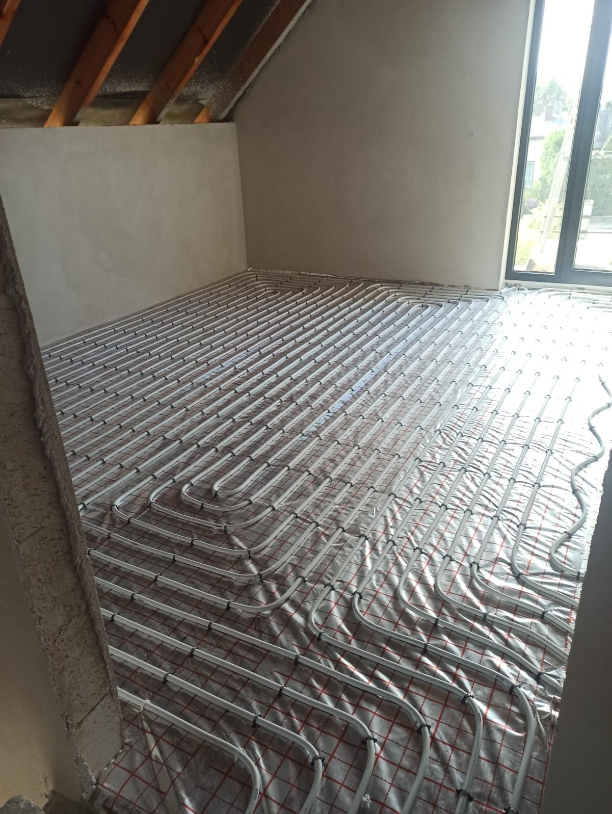 Białe rury ogrzewania podłogowego ułożone na folii z czerwoną kratą w surowym wnętrzu poddasza z oknem. Widoczne drewniane elementy konstrukcji dachu.