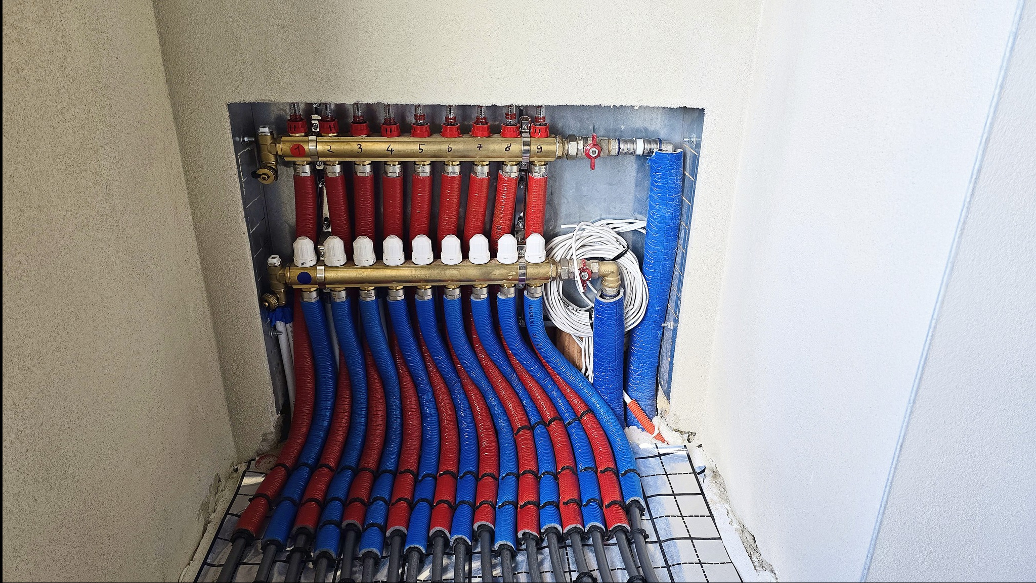 Rozdzielacz ogrzewania podłogowego z rurami w izolacji czerwono-niebieskiej, zamontowany w niszy ściennej. Widoczna siatka montażowa na podłodze i zawory regulacyjne.