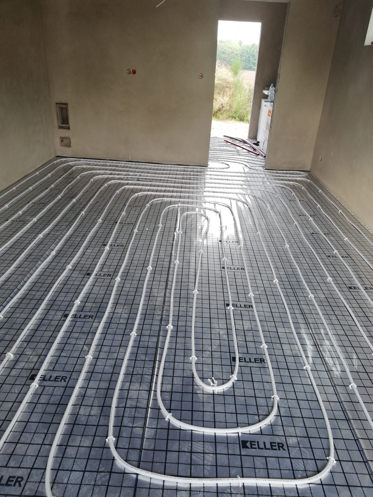 Ułożona instalacja ogrzewania podłogowego na folii z nadrukiem 'KELLER' w surowym wnętrzu, widok z góry, przygotowana do zalania posadzką.