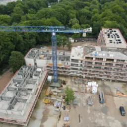 Budowa wielopiętrowego budynku z modułów, widok z góry. Na placu budowy stoi niebieski dźwig. Widoczne elementy konstrukcyjne i materiały budowlane.