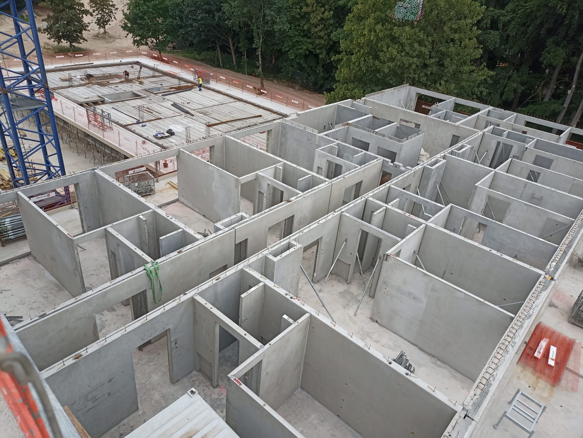 Wysoki kąt na budowę domu modułowego z prefabrykatów betonowych, widoczne ściany działowe i zewnętrzne, zbrojenia. Na drugim planie ekipa budowlana przy pracy.