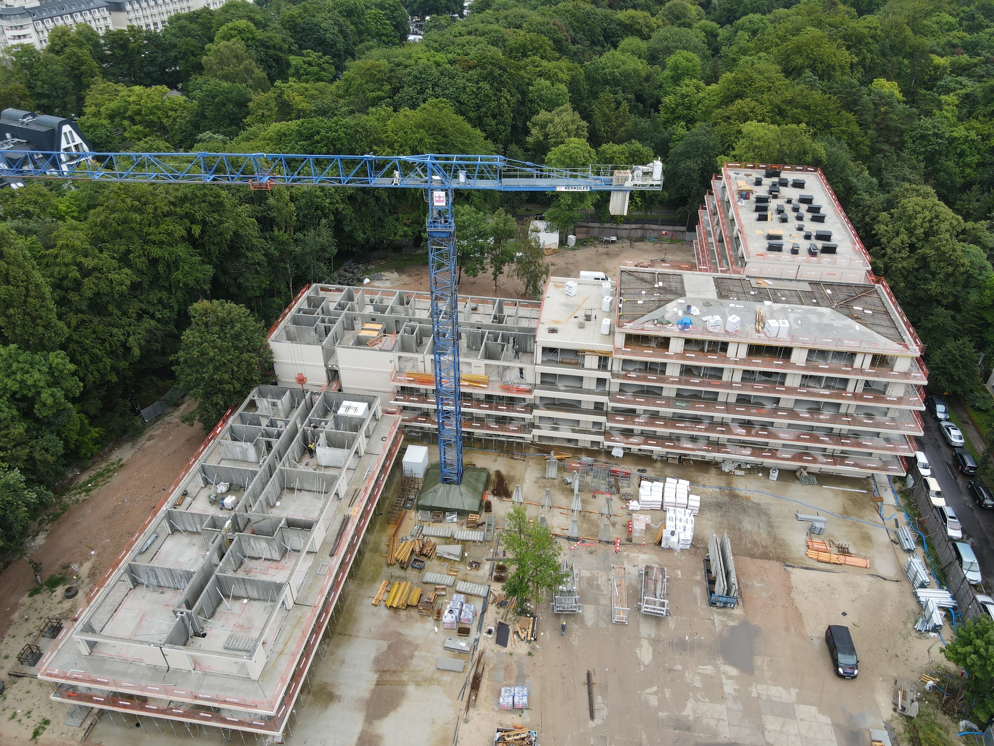 Budowa wielopiętrowego budynku z modułów, widok z góry. Na placu budowy stoi niebieski dźwig. Widoczne elementy konstrukcyjne i materiały budowlane.