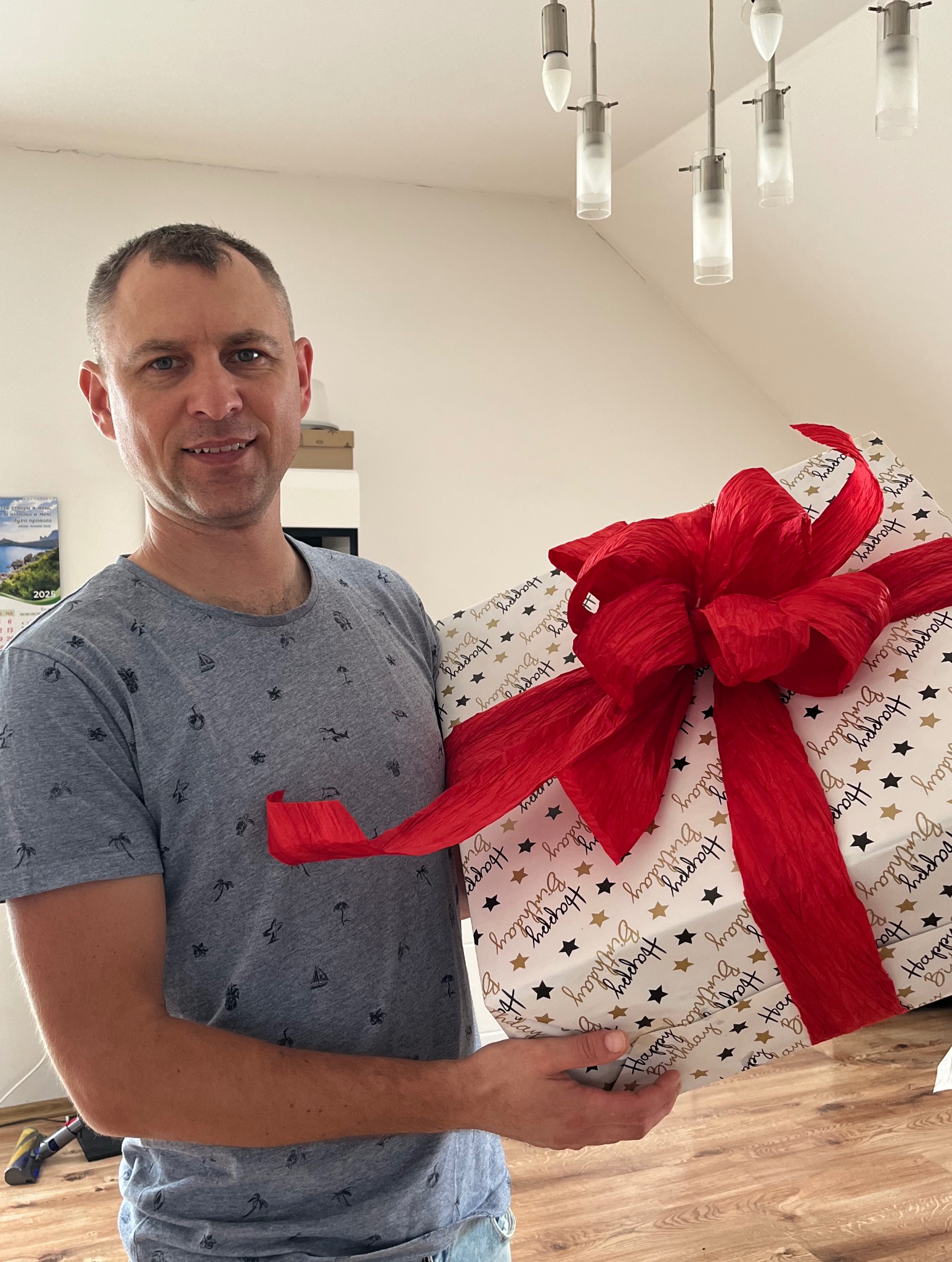 Uśmiechnięty mężczyzna trzyma duży prezent urodzinowy z czerwoną kokardą w jasnym wnętrzu, widoczne lampy sufitowe.