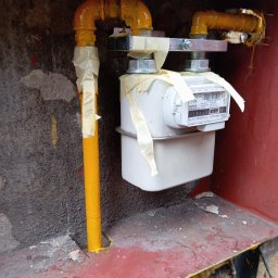 Gazownik-Gdańsk - Żółta instalacja gazowa z licznikiem, zabezpieczona taśmą malarską. Widoczne rury i mocowania. Ściana z szorstką fakturą w tle.