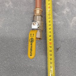 Gazownik-Gdańsk - Miedziana instalacja gazowa z zaworem KFA Armatura, pomiar długości za pomocą żółtej miarki zwijanej na szarym tle. Widoczne złącza i elementy instalacji.