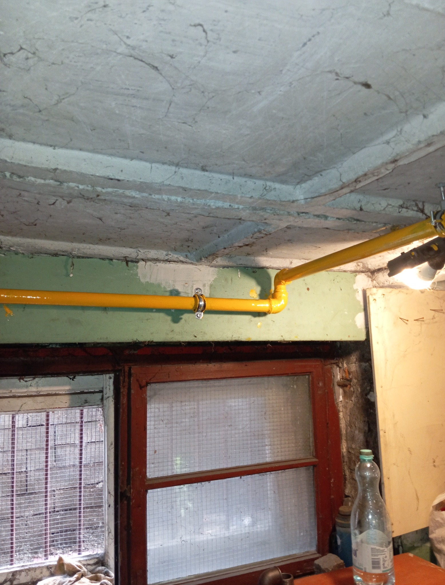 Żółta instalacja gazowa zamontowana w piwnicy, widoczna na tle okna z kratą i surowego sufitu. Rury prowadzone wzdłuż ściany, oświetlone lampą.