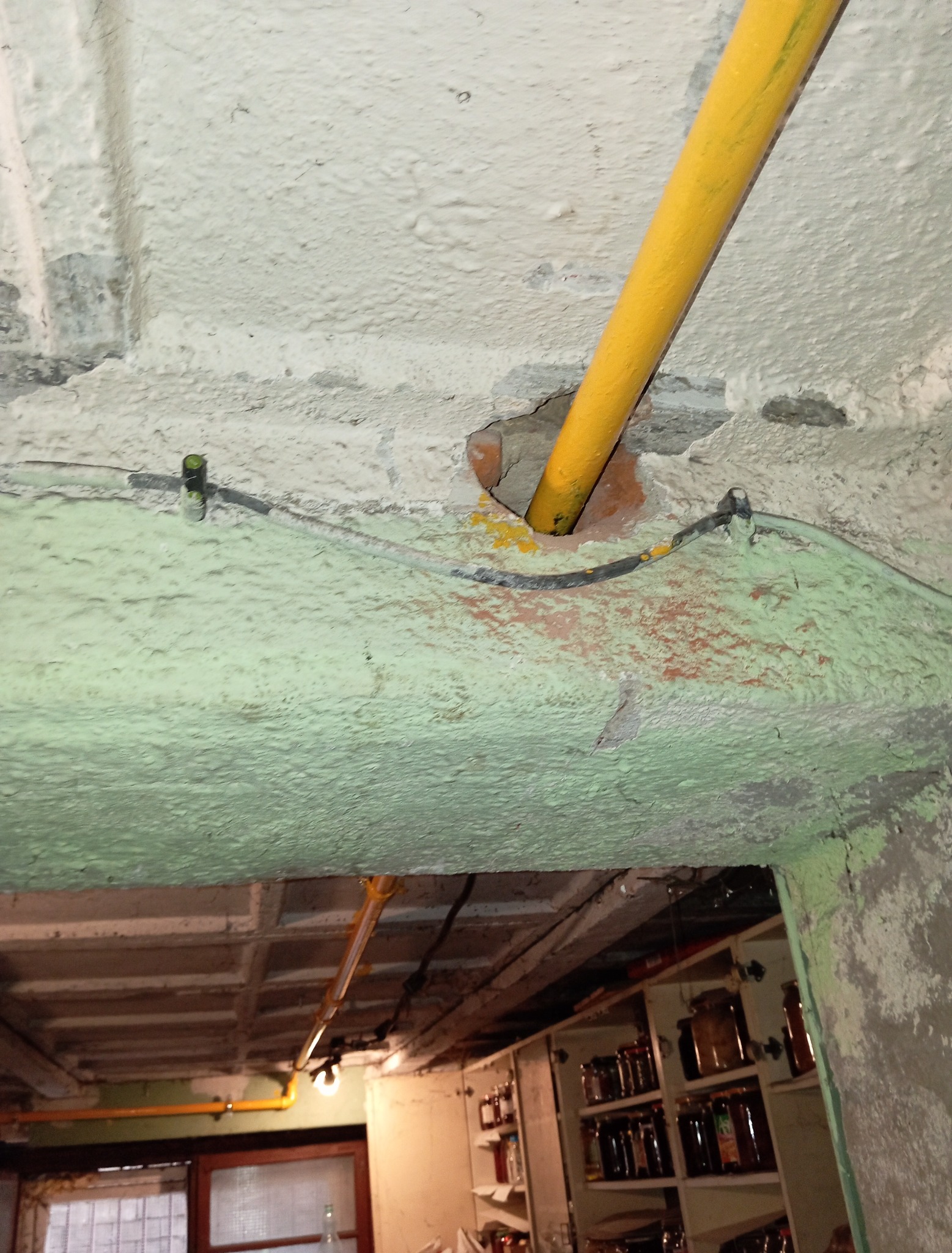 Żółta rura gazowa przechodzi przez dziurę w suficie w piwnicy. Widoczne instalacje i spiżarnia w tle. Nietypowe oświetlenie i surowe wykończenie.