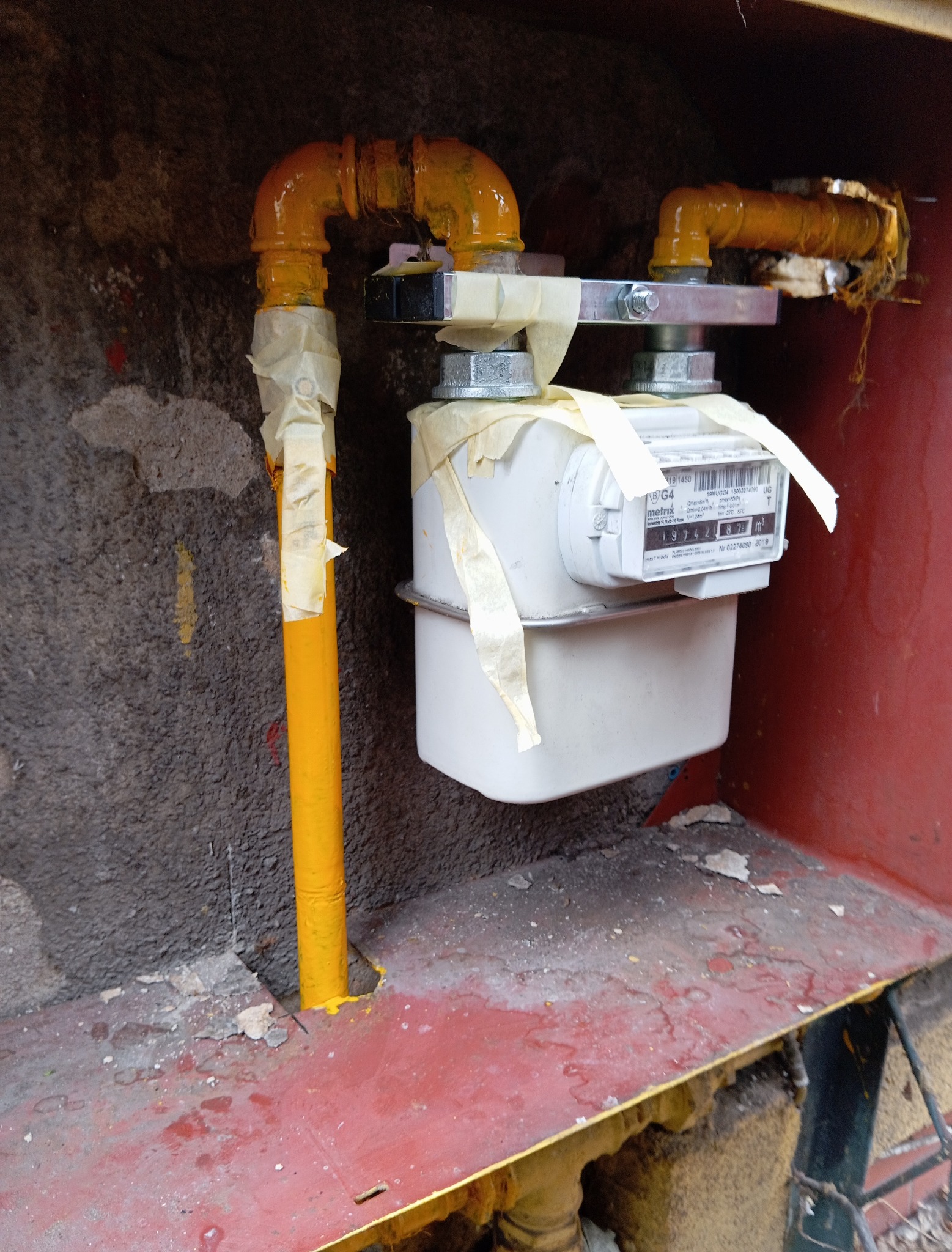 Żółta instalacja gazowa z licznikiem, zabezpieczona taśmą malarską. Widoczne rury i mocowania. Ściana z szorstką fakturą w tle.