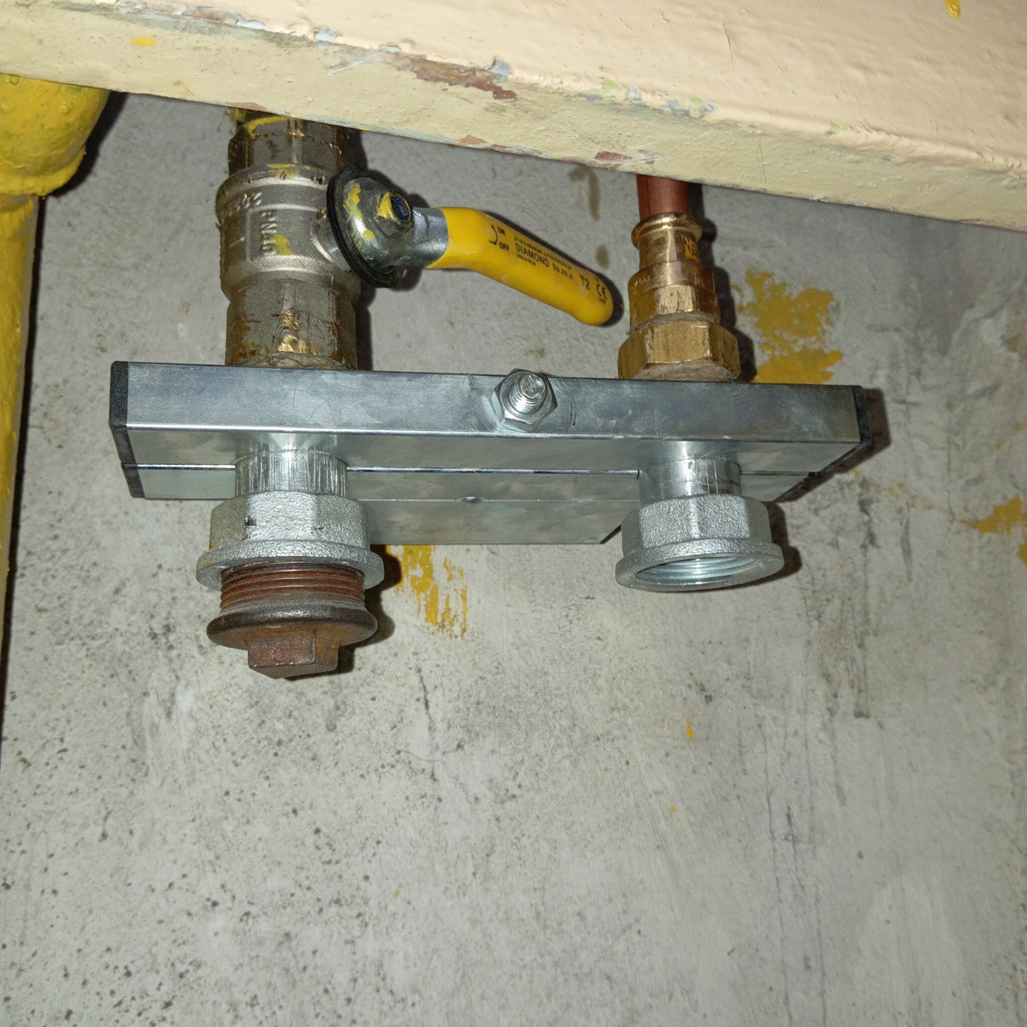 Szara, metalowa listwa rozdzielcza z zaworem gazowym z żółtą rączką, połączona z rurami, na tle betonowej ściany z żółtymi plamami.