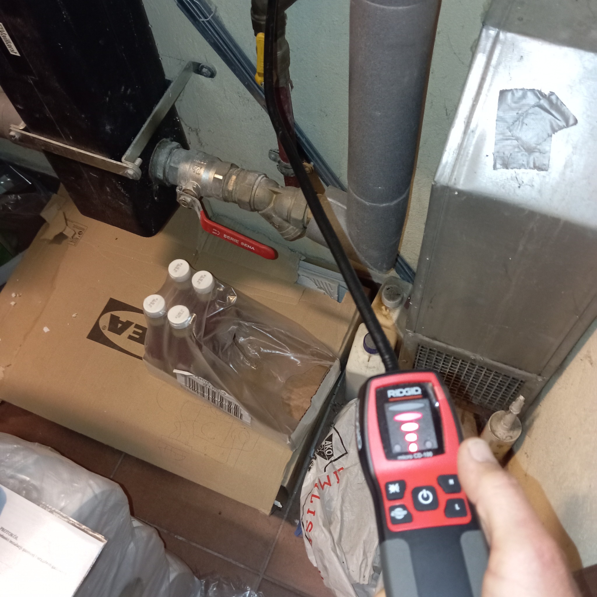 Kontrola szczelności instalacji gazowej detektorem RIDGID CD-100 w Gdańsku. Widoczny zawór, rury i karton IKEA. Nieostre ujęcie z bliska.