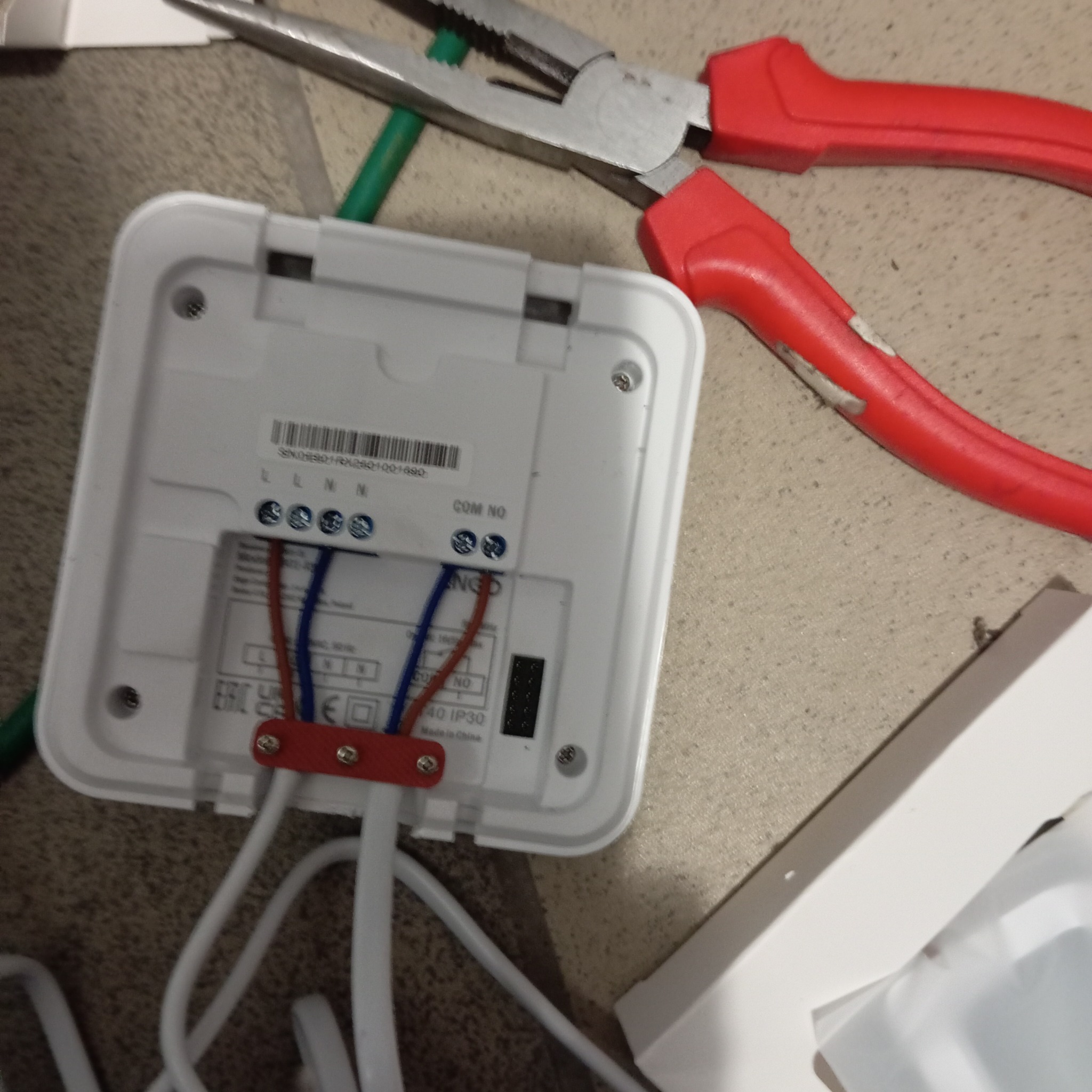 Podłączenie przewodów do białego panelu sterowania z użyciem czerwonych szczypiec. Widoczne śruby, oznaczenia L, N, COM NO oraz kable w izolacji.