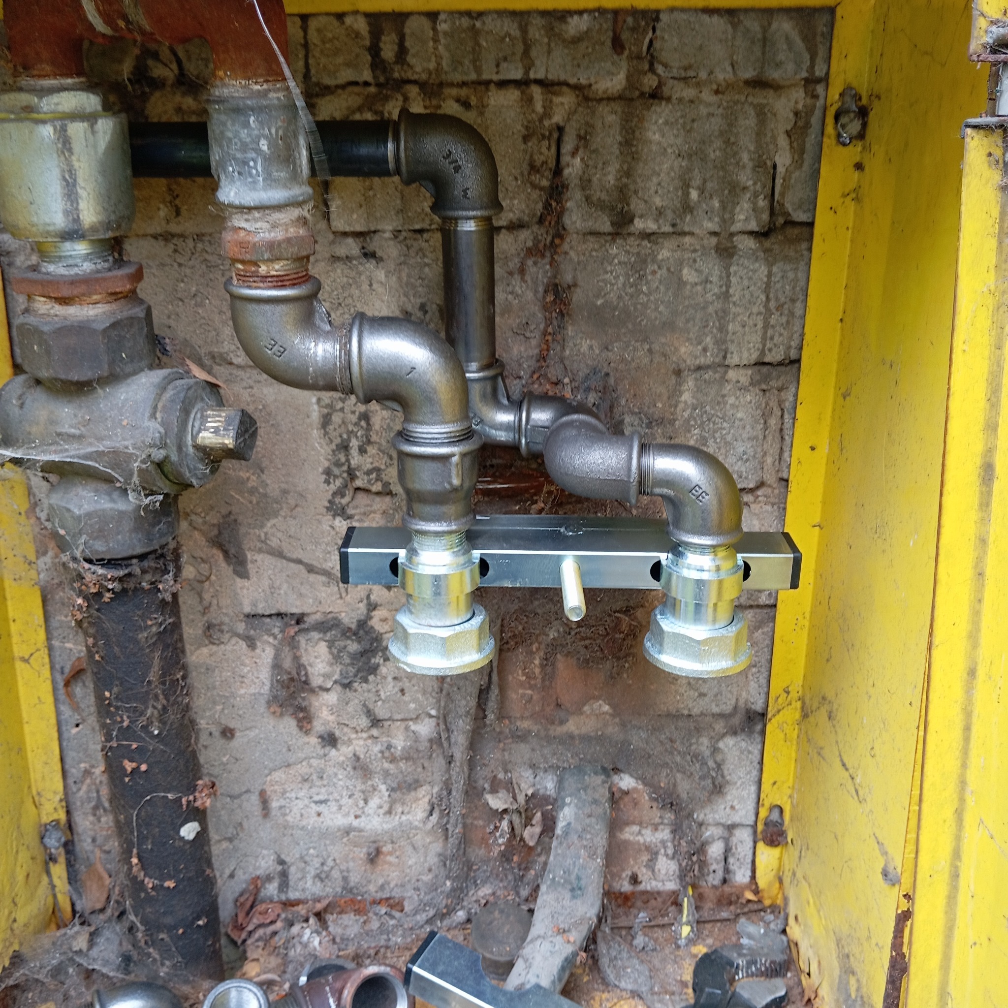 Złożona instalacja gazowa z metalowych rur i złączek, zamontowana na ścianie z cegieł w żółtej obudowie. Widoczne elementy instalacji i fragment ściany.