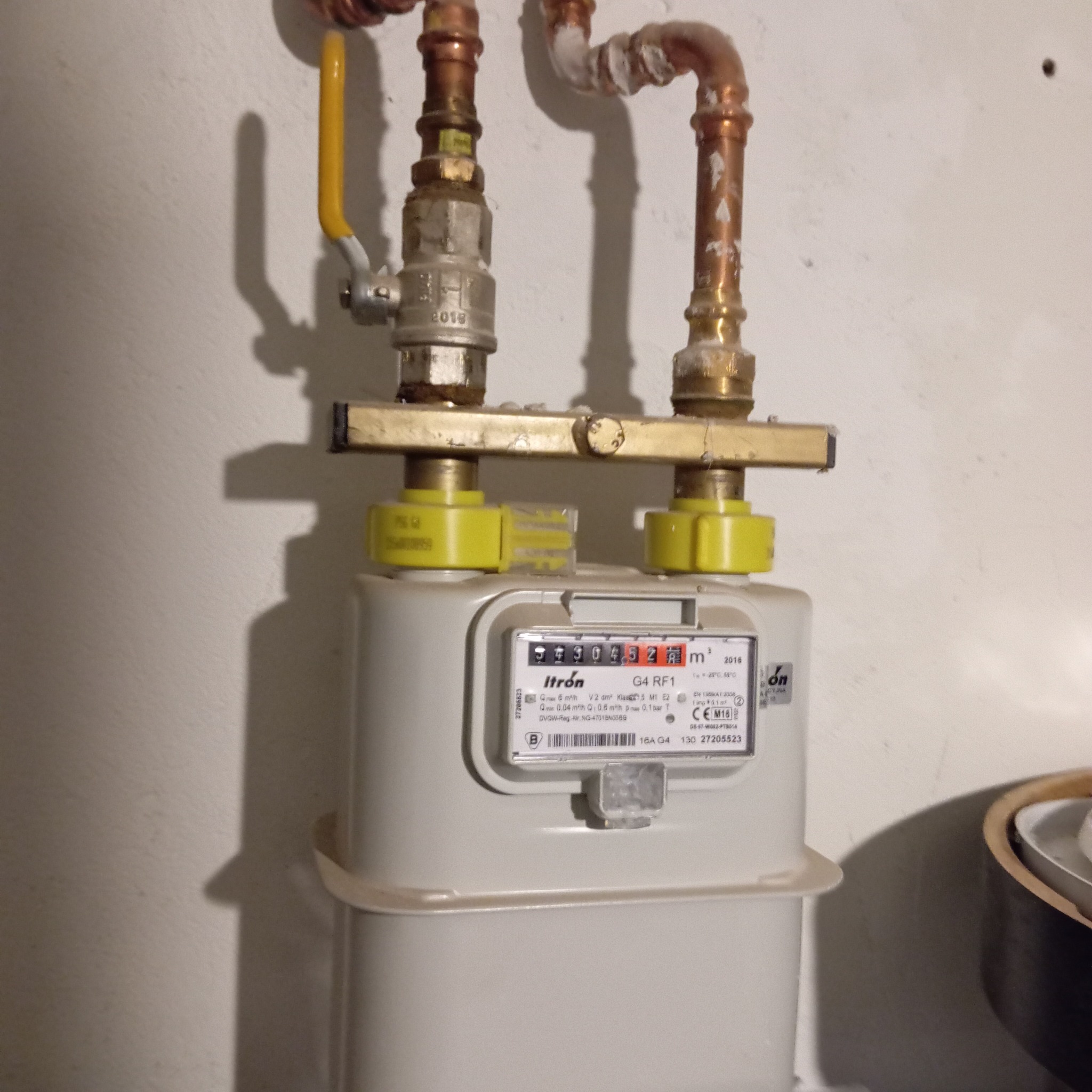 Zamontowany licznik gazu Itron G4 RF1 z żółtymi złączkami, rurami miedzianymi i zaworem. Widoczne oznaczenia i numery seryjne. Instalacja przy białej ścianie.