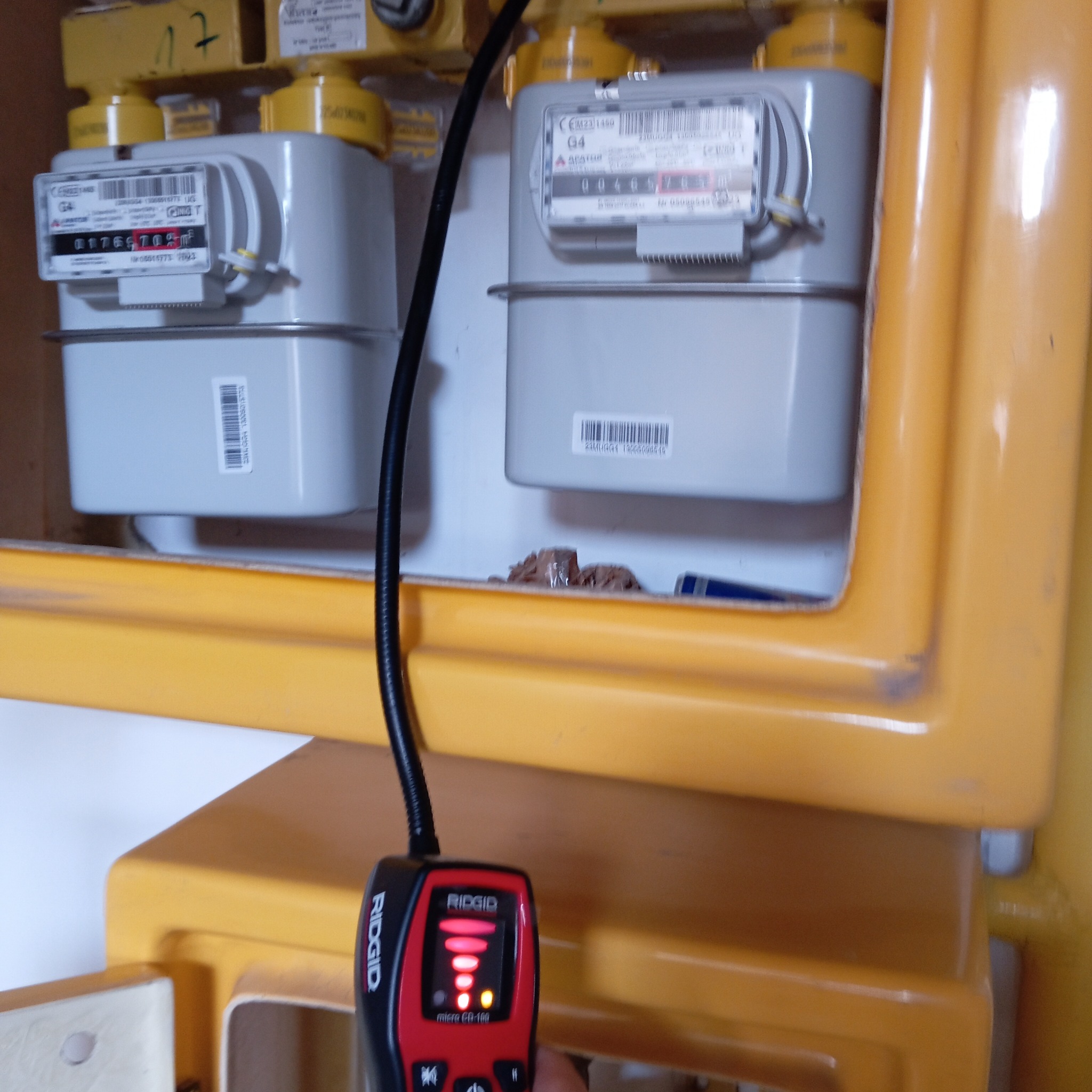 Dwa gazomierze w żółtej szafce, sprawdzane detektorem nieszczelności Ridgid, z widocznymi wskazaniami. Kontrastowe oświetlenie podkreśla detale.