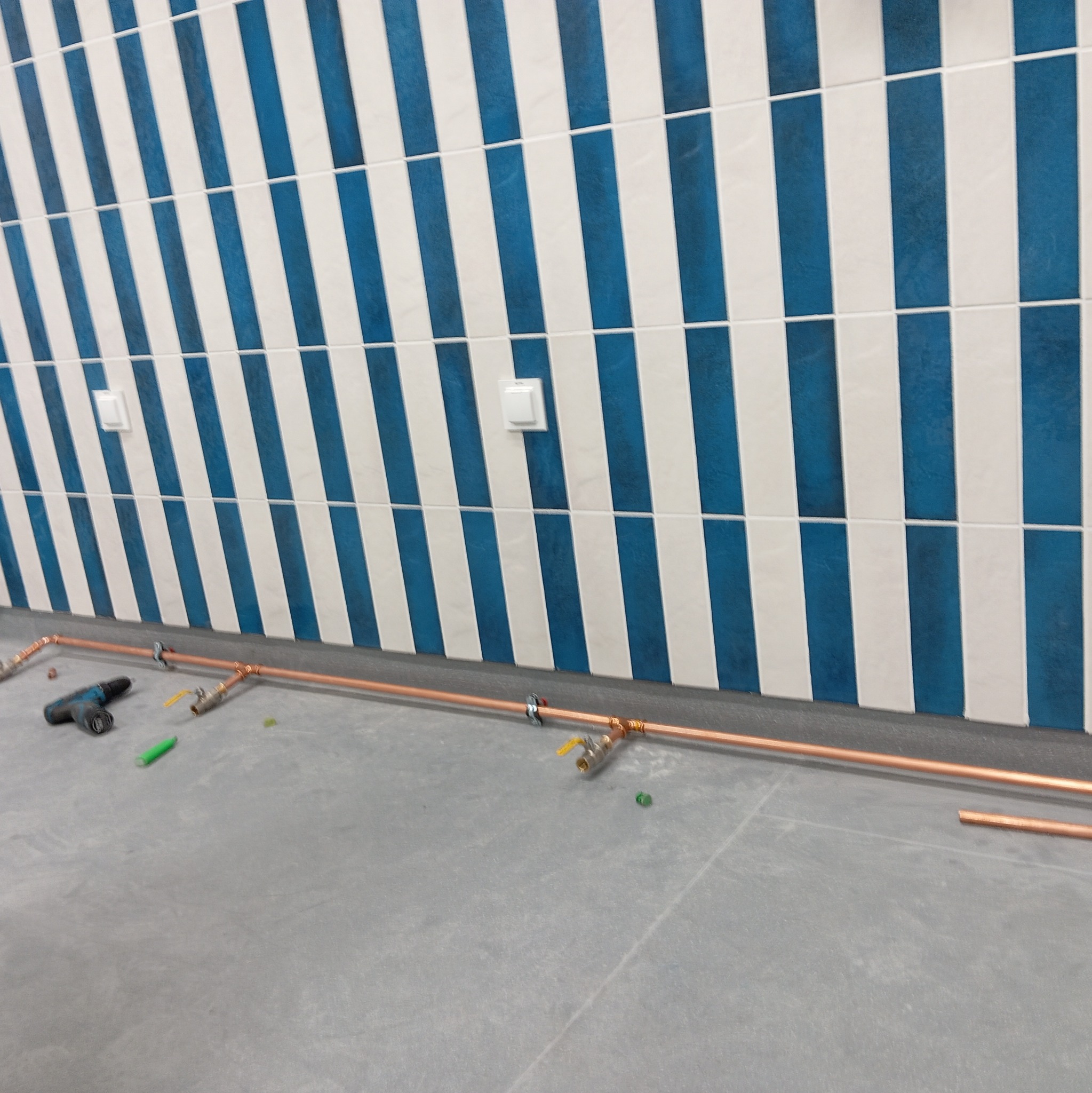 Miedziana instalacja gazowa w trakcie montażu, widoczne rury, zawory i narzędzia na tle ściany z biało-niebieskimi płytkami. Etap prac instalacyjnych.