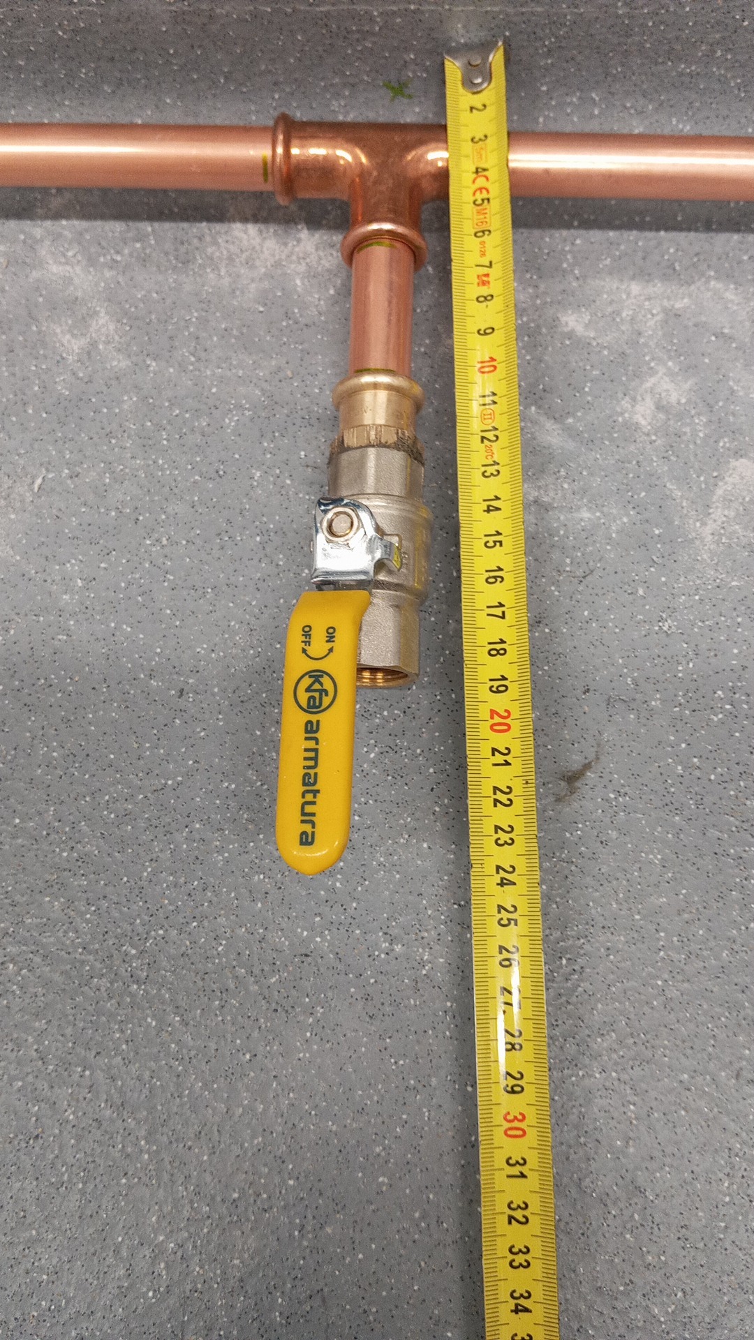 Miedziana instalacja gazowa z zaworem KFA Armatura, pomiar długości za pomocą żółtej miarki zwijanej na szarym tle. Widoczne złącza i elementy instalacji.