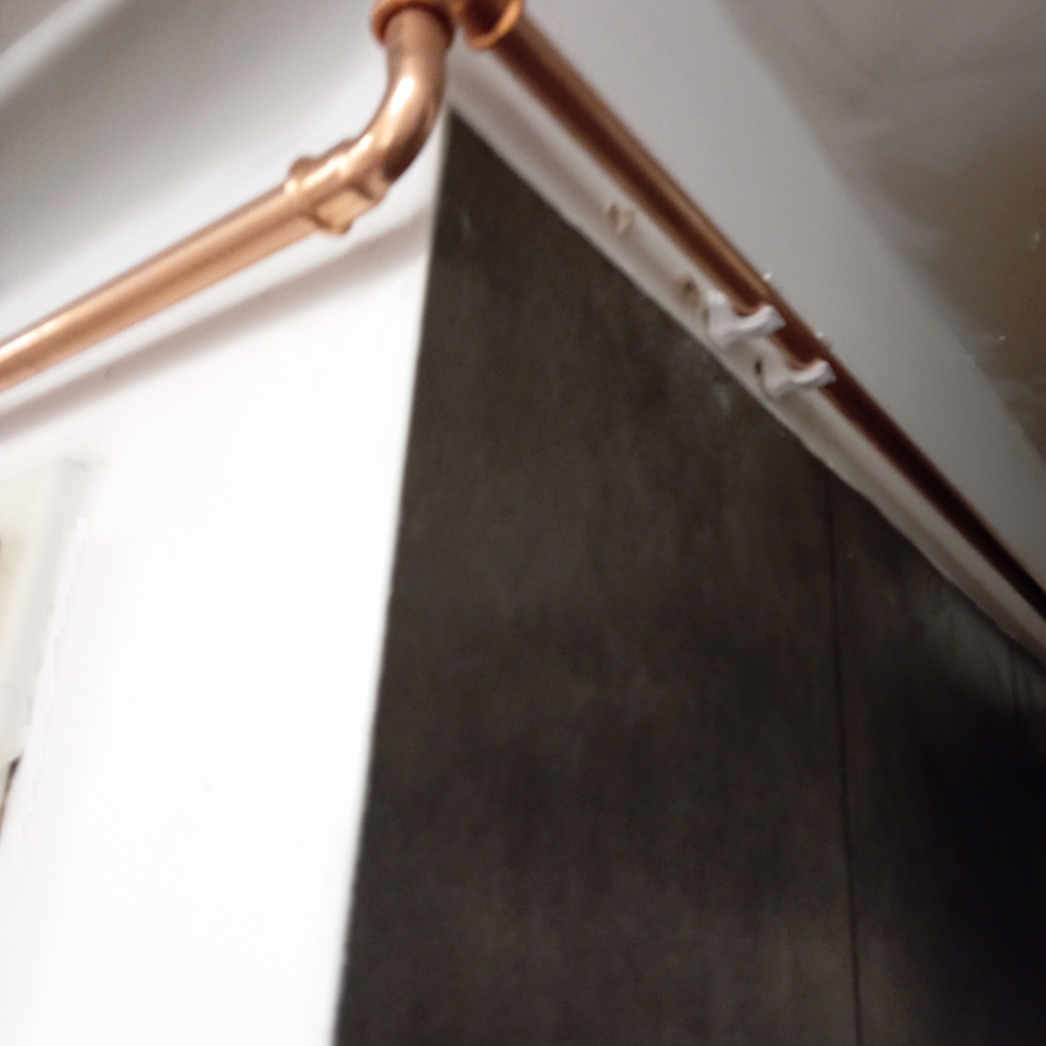 Miedziana rura instalacji gazowej, zamocowana do sufitu za pomocą białych uchwytów, na tle białej ściany i szarej płyty. Widok z dołu, ukośny kadr.