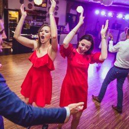 Michał Michalski Studio Fotowizualne - Rozbawieni goście w czerwonych sukienkach tańczą na parkiecie podczas imprezy. W tle oświetlenie sceniczne i bar. Dynamiczne ujęcie z perspektywy uczestnika zabawy.