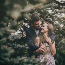 Michał Michalski Studio Fotowizualne - Uśmiechnięta para w objęciach na tle zielonych drzew iglastych. Kobieta w kwiecistej sukience, mężczyzna w marynarce. Romantyczna atmosfera, naturalne światło.