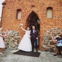 Michał Michalski Studio Fotowizualne - Para młoda wychodzi z kościoła obsypywana konfetti, w tle ceglana fasada i wiwatujący goście z tubami. Kadr z reportażu ślubnego.