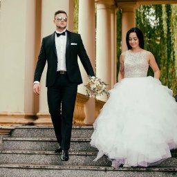 Michał Michalski Studio Fotowizualne - Elegancka para młoda schodzi po schodach, trzymając się za ręce. Pan młody w czarnym smokingu i okularach, panna młoda w białej sukni z bukietem. Kolumnada w tle.