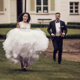 Michał Michalski Studio Fotowizualne - Uśmiechnięta panna młoda w sukni i trampkach ucieka po trawie, za nią biegnie pan młody w smokingu. W tle budynek z oknami i zielone krzewy.
