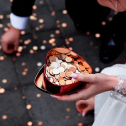 Panna młoda trzyma serce pełne monet, goście zbierają drobne z ziemi. Ujęcie z góry, widoczny fragment sukni ślubnej z koronkowymi rękawami.