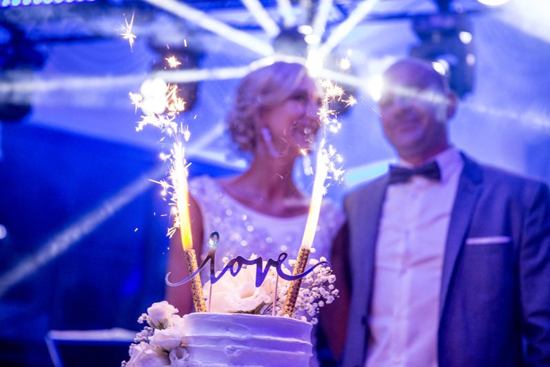 Uśmiechnięta para młoda za tortem weselnym z zimnymi ogniami i napisem 'love' w niebieskim oświetleniu podczas uroczystości.