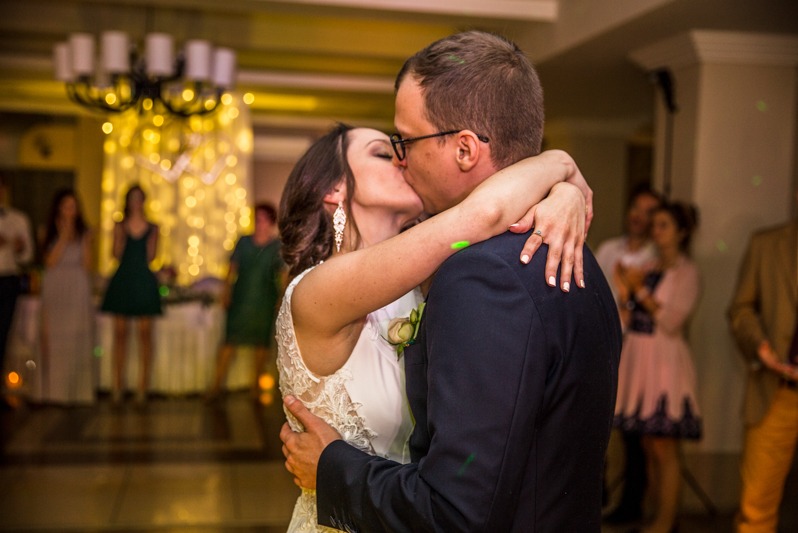 Pocałunek pary młodej podczas wesela, w tle goście i dekoracje świetlne. Panna młoda w koronkowej sukni, pan młody w ciemnym garniturze i okularach.