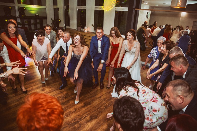 Wspólna zabawa weselna: goście w tańcu limbo pod sznurem rąk, para młoda w centrum, drewniany parkiet i dekoracyjne oświetlenie tworzą radosną atmosferę.